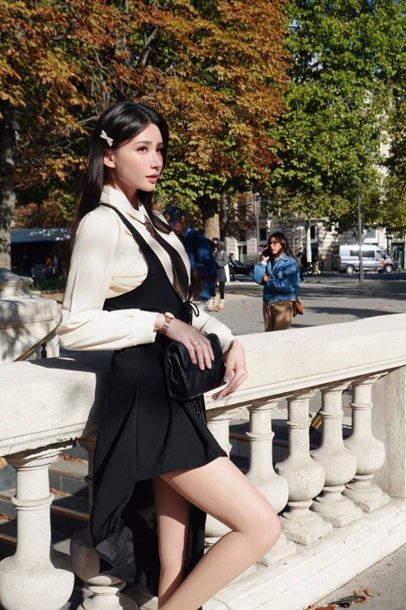 A young woman with long dark hair, wearing a white blouse and black dress with a small handbag, poses leaning against a stone balustrade in an outdoor urban setting with autumn trees.