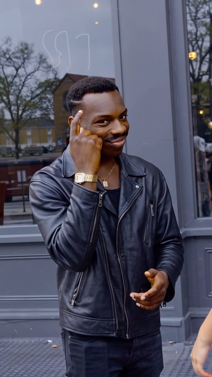 A stylish man in a black leather jacket, gold watch, and pendant necklace poses confidently with his finger to his temple outside a storefront. The image appears unrelated to the post about Pika Labs and memeheaven's GitHub fees, showing no connection to AI tools or financial platforms.