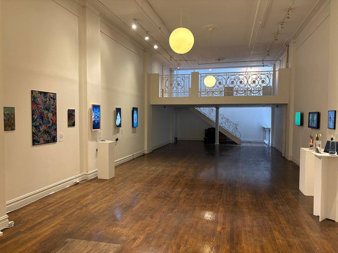An art gallery interior features wooden floors, white walls displaying colorful digital and abstract artworks, a central staircase with decorative railings, and large spherical pendant lights hanging from the high ceiling.