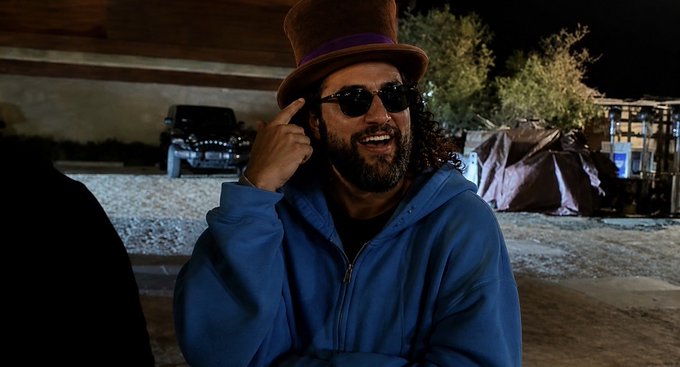 A bearded man wearing a blue hoodie, sunglasses, and a brown top hat sits outdoors at night, gesturing near his temple with parked vehicles visible behind him.