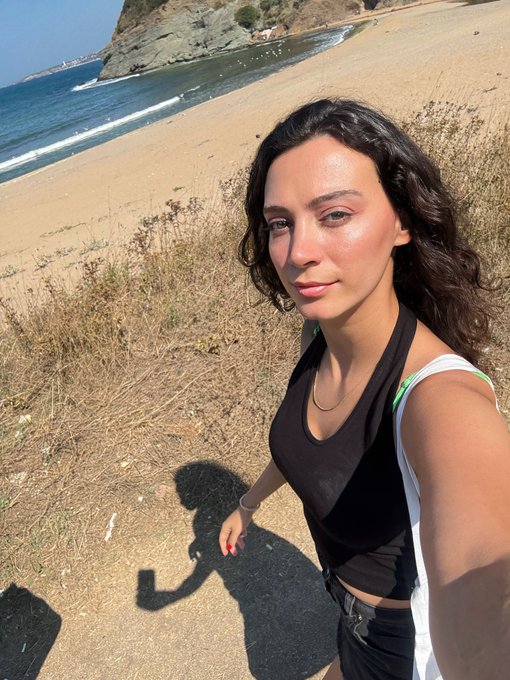 A woman with curly dark hair taking a selfie on a coastal trail, wearing a black halter top, with a sandy beach, ocean, and rocky cliffs in the background.