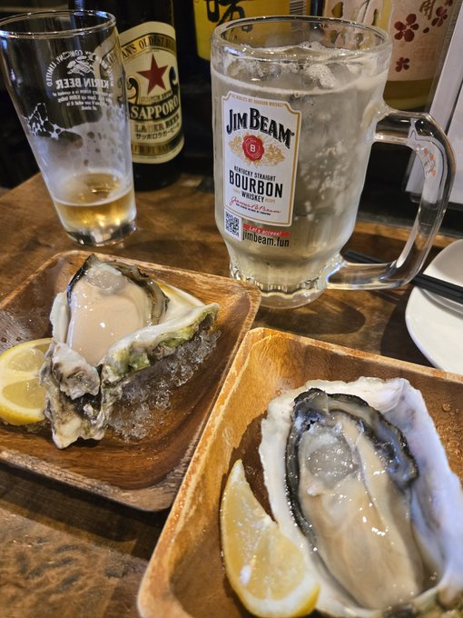 Two oysters on ice with lemon wedges served on wooden plates, accompanied by a glass of Jim Beam bourbon and a bottle of Sapporo beer in the background.