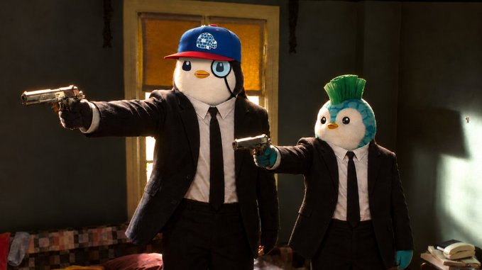 Two plush mascot characters, one resembling a penguin with a cap and the other a bird with a green mohawk, dressed in black suits and pointing guns in a dimly lit room, evoking a Pulp Fiction-esque scene.
