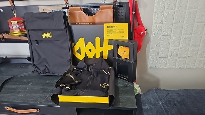A collection of Binance-themed merchandise gifts is displayed on a black surface, including a black backpack with yellow "BNB" lettering, a pair of black "Binance" branded gloves, a yellow "HODL" sign, a boxed yellow figurine, and an information card, all coordinating with the post's announcement of receiving premium gifts from the Binance TR team.