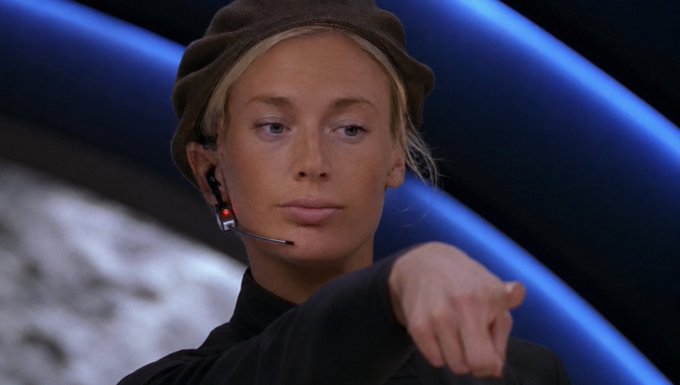A woman wearing a headset and dark cap is captured in a moment of intense focus, pointing her finger forward with a serious expression, as if she is about to initiate an action or "push the button." Her determined gaze and decisive gesture convey the urgency of the moment, fitting the prompt's call to action.