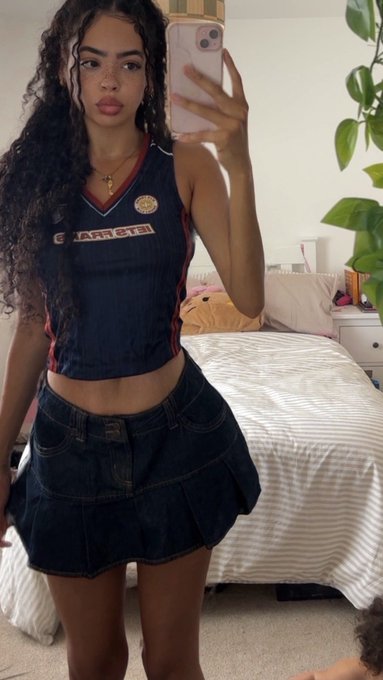 A young woman with long curly dark hair takes a mirror selfie in a bedroom, wearing a dark blue 'ETTERS' (Estrela) sports jersey and a dark denim skirt.