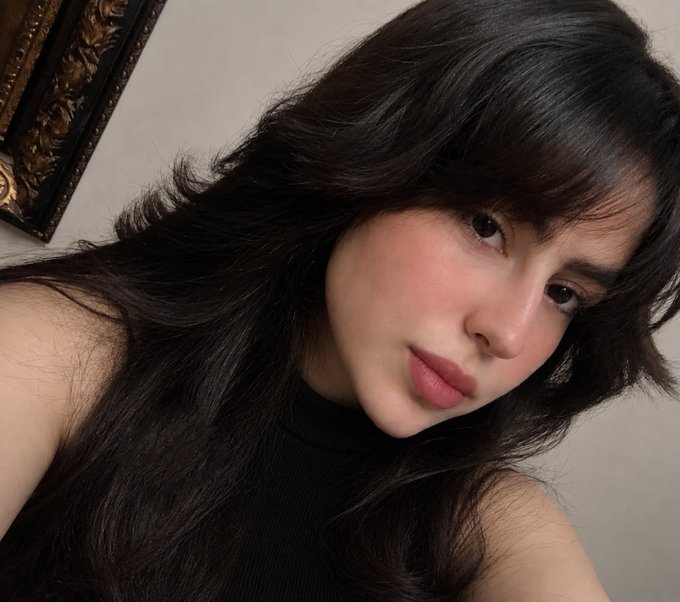 A close-up selfie shows a young woman with long dark hair, bangs, and pink lips, wearing a black sleeveless top.