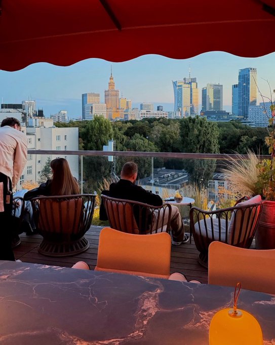 A rooftop bar view under a red umbrella overlooking the Warsaw skyline, including the Palace of Culture and Science, with people seated in chairs.