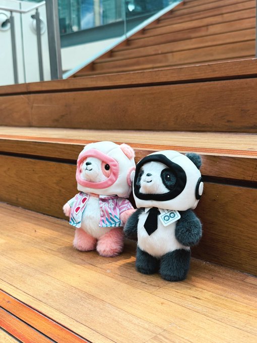 Two small plush toys—one dressed in a pink astronaut-like suit with a patterned jacket, and the other a panda in a suit and tie—sitting on wooden steps.