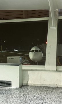 A view through an airport terminal window showing an airplane parked at a gate at night, with a luggage cart visible on the tarmac.