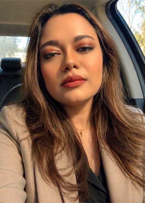 Close-up selfie portrait of a young woman with long brown hair wearing a tan blazer, sitting in a car with trees visible through the window behind her.