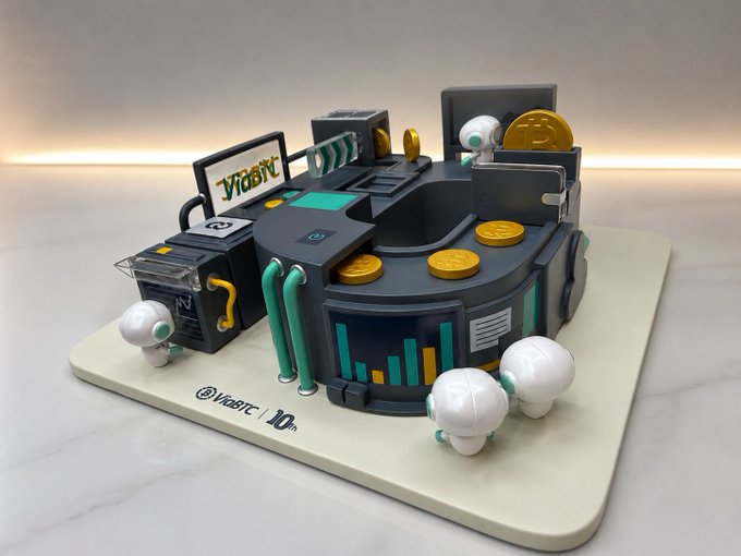 The image features a unique cake designed like a factory or office space with various elements such as desks, chairs, and computer monitors. The cake has been created using Lego pieces, giving it an industrial and creative appearance. The cake is placed on a white surface, which contrasts nicely with its colorful design. The post accompanying the image congratulates @ViaBTC for ten years of supporting Kaspa mining and mentions that the cake was made in their honor.