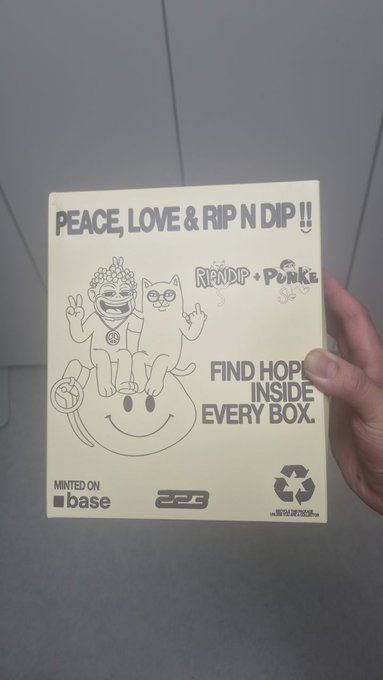 A hand holding a cream-colored 'PEACE, LOVE & RIP N DIP!!' RIPNDIP x PUNKE collaboration box, minted on Base blockchain, with text 'FIND HOPE INSIDE EVERY BOX.'