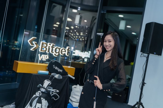 A smiling woman in a black dress poses with a golf club beside a "Bitget VIP" sign at the AWS x Bitget VIP Exclusive Event, where a branded golf bag and equipment are displayed. The setting suggests a networking or hospitality moment from the event, complementing the post's focus on the successful conclusion of the VIP gathering.