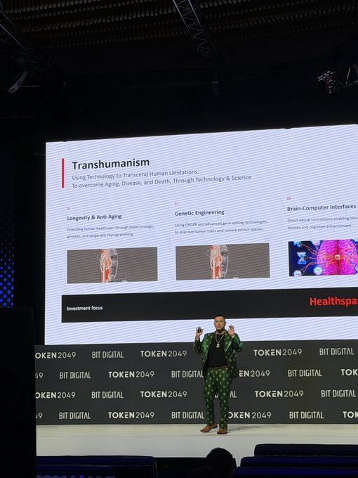 A man in a green patterned suit presents on stage at the TOKEN2049 BIT DIGITAL conference, with a slide behind him about 'Transhumanism' covering anti-aging, genetic engineering, and brain-computer interfaces.
