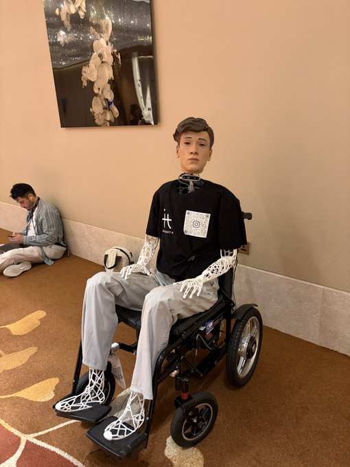 A humanoid robot with a realistic human-like face sitting in a wheelchair, wearing a black t-shirt with a QR code and light gray pants, with exposed mechanical skeletal hands, positioned in what appears to be a hotel hallway.
