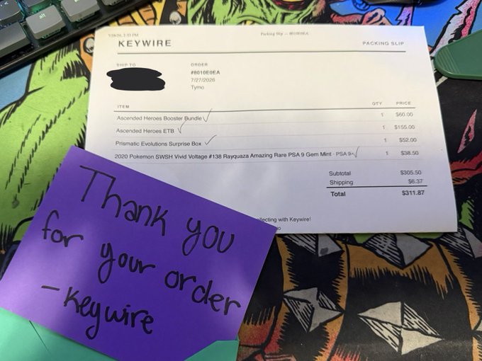 A Keywire packing slip and handwritten purple thank-you note from "Keywire" rest on a colorful Pokémon-themed desk mat. The receipt shows an order from "Tymo" on July 28, 2026, for an Ascended Heroes Booster Bundle ($60), Ascended Heroes ETB ($155), Prismatic Evolutions Surprise Box ($52), and a 2020 Pokémon SWSH Vivid Voltage PSA 9 Rayquaza ($38.50)—totaling $311.87. The image illustrates a satisfied customer's order alongside a sweet personal note, perfectly matching the post's celebration of happy Keywire customers.