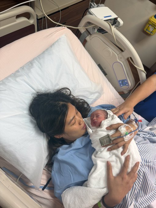 A new mother in a hospital bed holds her swaddled newborn, who still has a hospital ID tag attached to their tiny wrist. The intimate post-birth moment humorously celebrates the baby's faster "mainnet launch" compared to the developers still working on BULK.