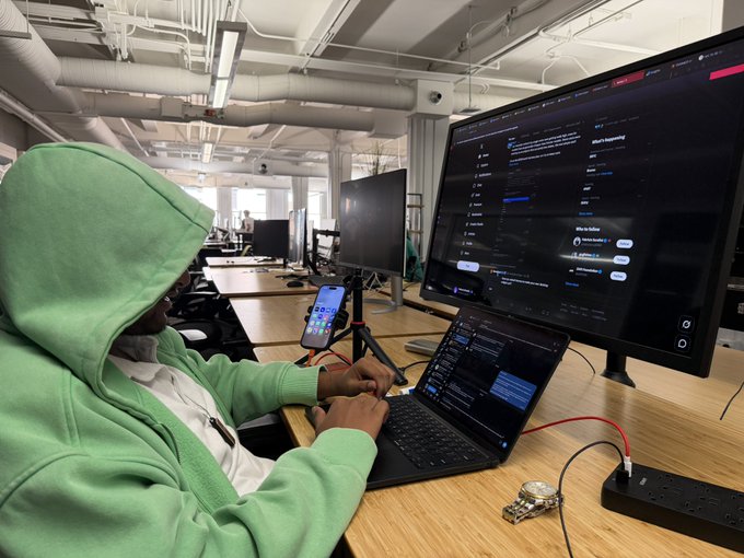 A person wearing a green hoodie working at a desk in an industrial-style office, using a laptop connected to a large monitor displaying a dark-themed messaging application interface.