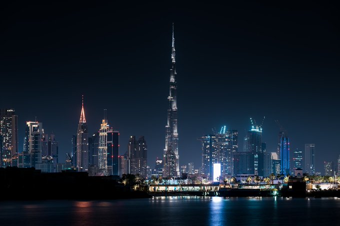 A nighttime cityscape of Dubai showcases the illuminated Burj Khalifa towering over surrounding skyscrapers, with their lights reflecting on the water in the foreground.