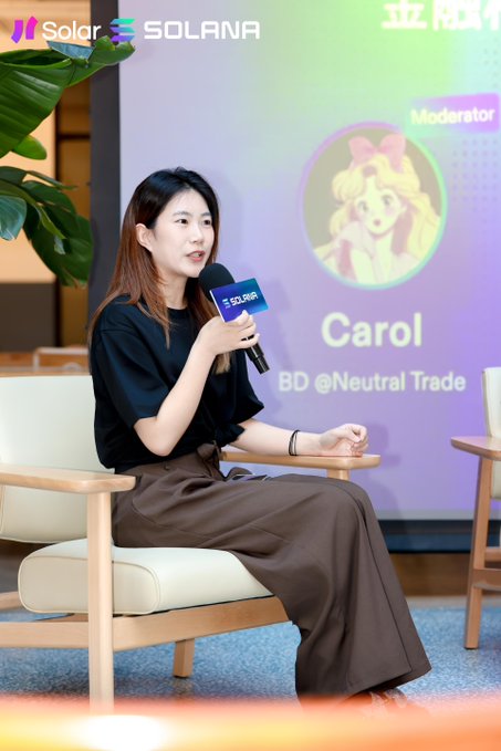 A woman in black sits in a chair speaking into a Solana-branded microphone at a Solana event; the screen behind her shows 'Moderator Carol, BD @Neutral Trade.'
