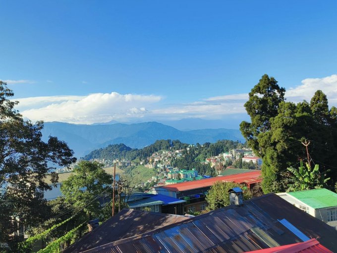 A scenic landscape view featuring a town with colorful rooftops nestled in lush green hills and mountains, with a body of water visible on the left, all under a clear blue sky.