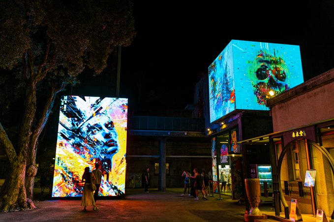 A nighttime urban scene with large illuminated digital art displays featuring colorful faces, where pedestrians walk by the glowing installations near some storefronts.