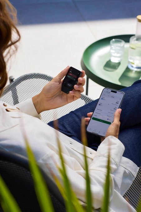 A person lounges outdoors holding a Trezor hardware wallet in one hand while checking a smartphone app, likely their crypto portfolio, in the other—visually reinforcing the endorsement of Trezor as a secure, user-friendly way to manage and store Bitcoin and other cryptocurrencies.