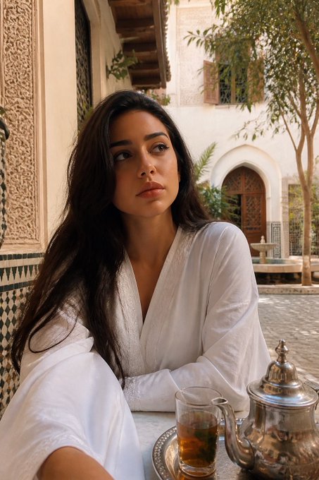 A woman with long dark hair wearing a white robe sits at a table with a glass of mint tea and an ornate silver teapot, set in a Moroccan-style courtyard.