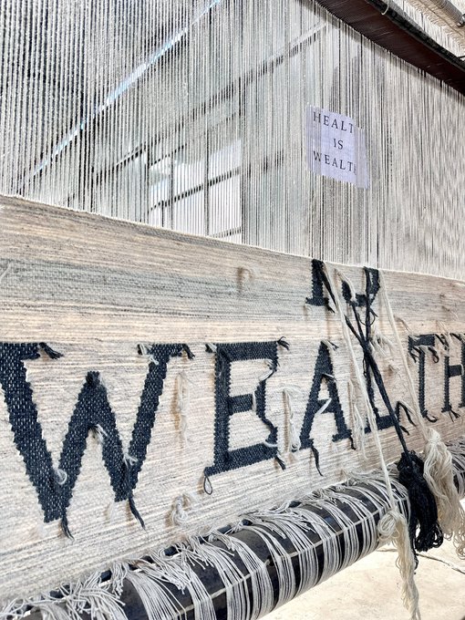 A weaving loom in progress displays the word 'WEALTH' being woven in large black letters onto the fabric, with a small printed sign hanging above that reads 'HEALTH IS WEALTH'.