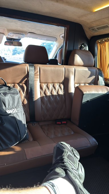 The interior of a vehicle features brown quilted leather seats, a black backpack on the floor, and a person's foot wearing a sneaker resting in the foreground.