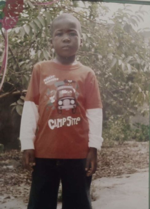 A young boy stands outdoors wearing an orange layered shirt featuring a vehicle graphic and the text "Campsite" along with "Honest Earners." His serious, bald appearance and innocent expression contrast with the post's claim that he is being unfairly billed for a wig, highlighting the absurdity of the situation.