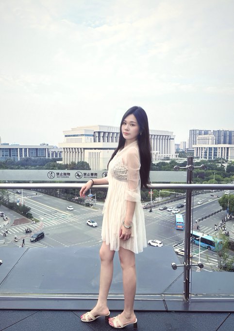 A young woman in a cream-colored dress standing on a balcony overlooking a city street with traffic and a large stadium-like building in the background.