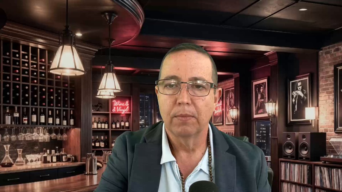 The image features a man sitting at a bar, wearing glasses and a suit jacket. He appears to be looking directly into the camera with an expression of concern or worry. The background includes several wine bottles displayed behind him, along with a few other people in the scene. There is also a neon sign that reads "Cardano".