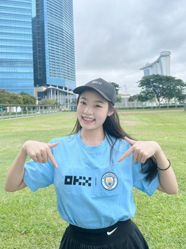 The image features a young woman wearing a blue shirt with "okx" written on it, standing outdoors in front of a building. She has her hands up in the air, pointing at herself as if posing for a picture. The background includes a grassy field and some trees, creating an outdoor setting. The post seems to be related to OKX, possibly discussing their business or promoting a product.