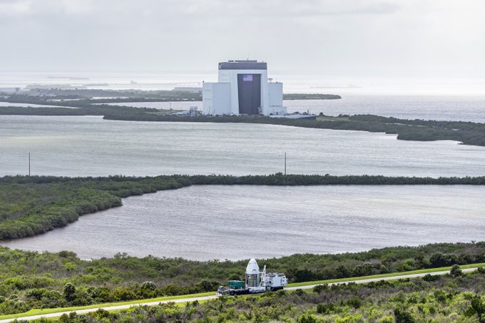 An aerial view shows a large white NASA building with an American flag situated on a coastline, with waterways and marshland in the foreground.