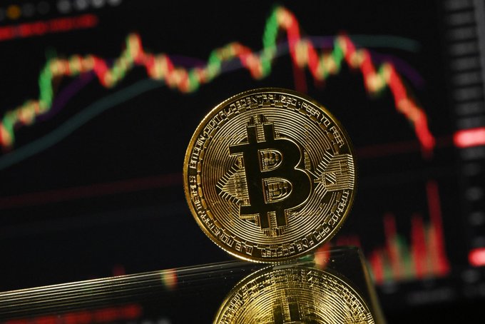 Photograph of a physical gold-colored Bitcoin token standing upright with a blurred stock market chart showing red and green candlestick patterns in the background.