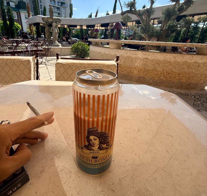 A tall, orange-striped "RE DI ROMA" beer can sits on a sunlit café table, with a hand holding a cigarette beside it and a Mediterranean terrace scene in the background. The image humorously pairs the "Re di Roma" (King of Rome) branding with the post's bullish, confident crypto commentary about Monadcards and Kuru, suggesting a relaxed "king-sized" victory moment amid the hype.