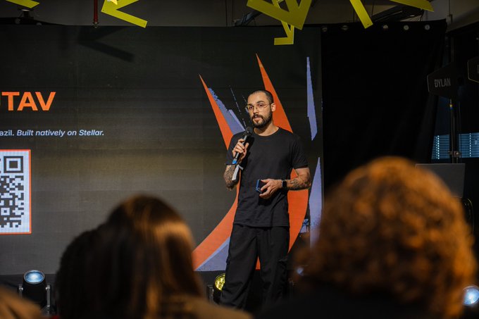 A man with glasses and tattoos speaking into a microphone on stage at a presentation, with a screen behind him reading 'TAV' and 'Built natively on Stellar' alongside a QR code.
