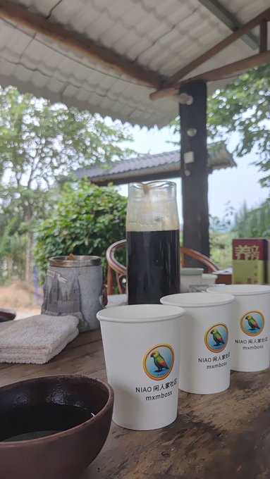 A rustic outdoor scene featuring a wooden table with paper cups branded 'NIAO 南人果吧 m&mboss' and a bottle of dark liquid, set against a backdrop of a covered patio and green foliage.