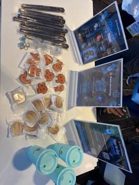 The image shows an assortment of PancakeSwap merchandise, including plush pancake toys, blue cylindrical items (likely water bottles or containers), lanyards, and framed promotional posters featuring PancakeSwap branding and cartoon mascots, all laid out on a table at the "kbw" (Korea Blockchain Week) event booth. The caption connects to the post by showcasing the exclusive PancakeSwap merch being promoted, where attendees can visit the booth to spin for these branded giveaways.