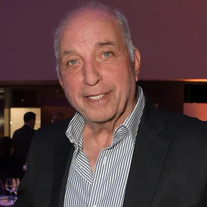 A headshot of an older man with gray hair and blue eyes, wearing a dark jacket over a striped shirt, smiling at the camera.