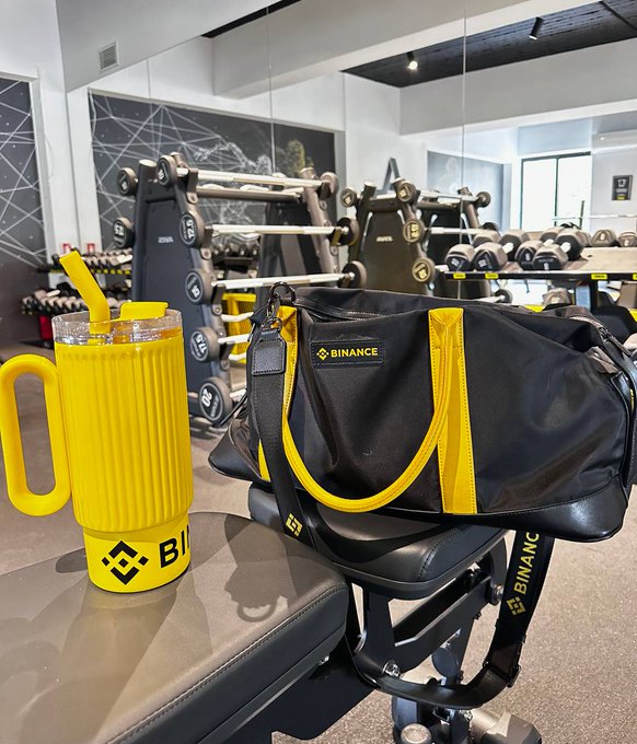 A yellow "BINANCE" branded tumbler and matching duffel bag rest on gym equipment in a well-equipped fitness center. The branded accessories perfectly complement the morning workout check-in theme, showing crypto-fitness lifestyle motivation.