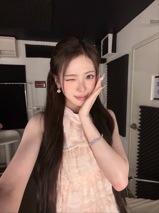 Same image as Image 4: a woman with long dark hair winks at the camera, resting her cheek on her hand, wearing a light pink lace top in a studio setting.