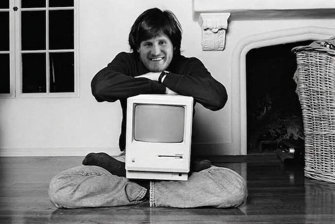 A black-and-white photo of a smiling young man leaning on a vintage Apple Macintosh computer while sitting cross-legged on a floor.