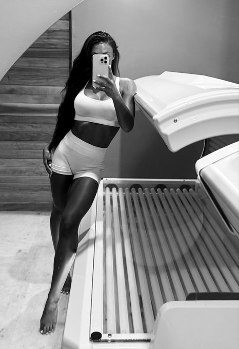 A black and white photo of a woman taking a mirror selfie, wearing a white sports bra and shorts next to a tanning bed.