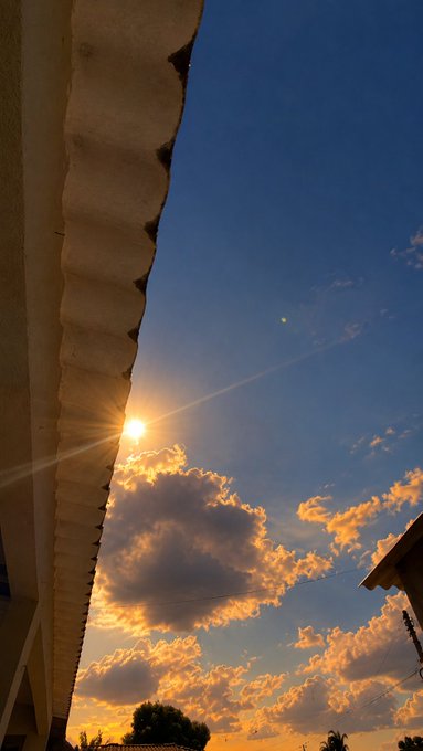 A sunset photo looking up past the edge of a roof, showing the sun creating a lens flare in a blue sky with scattered golden clouds.