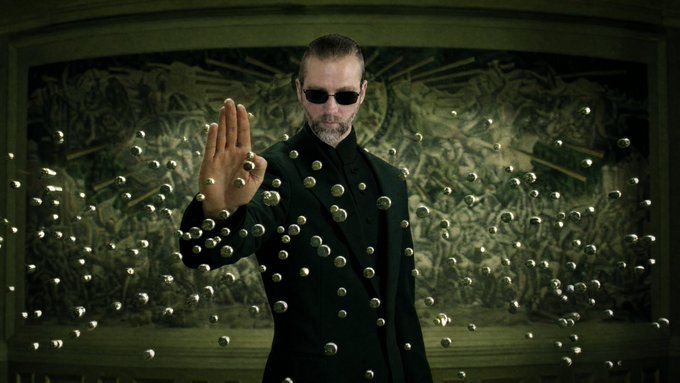 A scene from The Matrix featuring the character Agent Smith wearing a black suit and sunglasses, holding up his hand to stop bullets that are frozen mid-air around him.