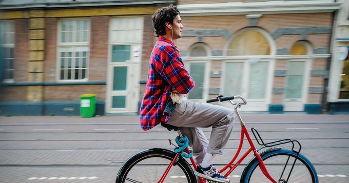 A young man in a red plaid shirt casually rides a red bicycle down a street lined with classic European brick buildings, moving at a relaxed pace while looking forward with a slight smile. The leisurely, carefree cycling parallels the unhurried, confident approach of $TIBBIR holders contrasted against the methodical, calculated TWAP strategy employed by large institutions.