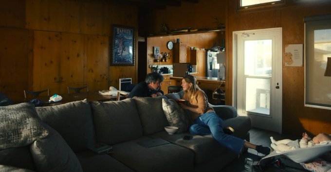 A dimly lit, wood-paneled living room scene where a man and woman sit close together on a large sectional sofa, leaning toward each other intimately, with a kitchen and a door visible in the background.